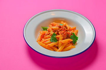 Plate of penne pasta with tomato sauce served in a white plate over bright pink background. Copy space banner.