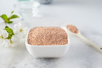 Ceramic bowl with red clay powder, for homemade facial and body mask or scrub and fresh sprig of flowering cherry on white background. Spa and bodycare concept.