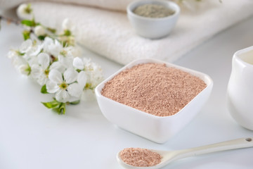Fototapeta na wymiar Ceramic bowl with red clay powder, ingredients for homemade facial and body mask or scrub and fresh sprig of flowering cherry on white background. Spa and bodycare concept.