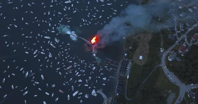 Unique revealing, high altitude, top down aerial drone footage of small motor boats and sail boats gathering around and witnessing the biggest bonfire event on Earth at Alesund, Norway at dusk.