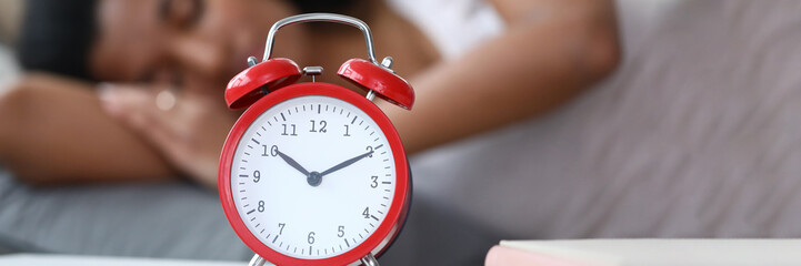 Close-up of red mechanical alarm clock standing on bedside table showing ten minutes to eleven. Young latino woman enjoying sweet dream on background. Wake up concept