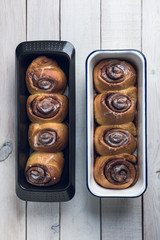 Fresh homemade cinnamon rolls on two baking pans, black and white, over a white rustic wooden table. Top view.