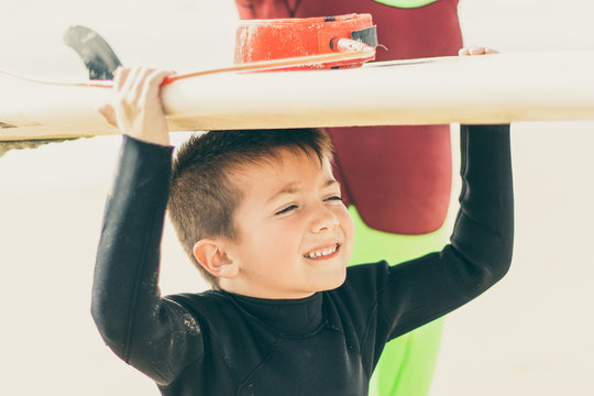 Cute Little Boy Holding Surfboard. Adorable Smiling Child In Wetsuit Holding Surfboard And Looking Aside On Sandy Beach. Surfing Concept