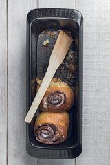Black baking pan with two fresh homemade cinnamon rolls and a spatula, over a white rustic wooden table. Top view
