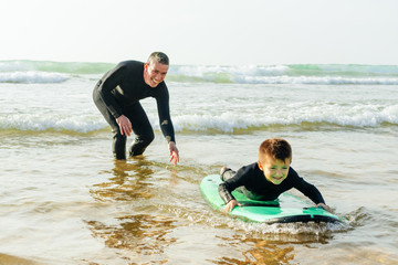 Father teaching son surfing in ocean. Happy father helping little son in wetsuit lying and swimming on surfboard on waves. Surfing concept