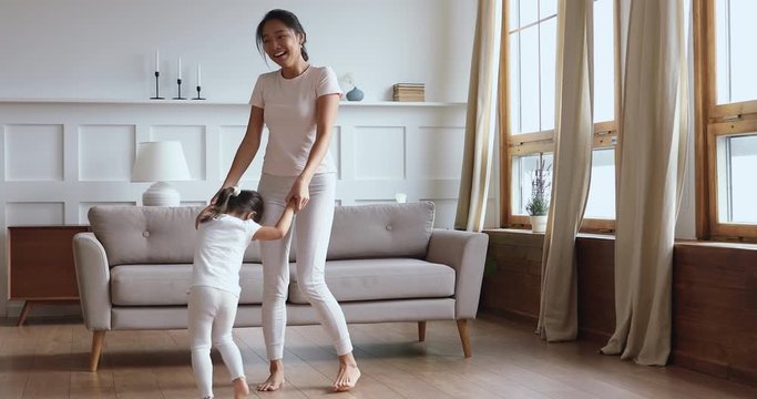Full length overjoyed young asian woman holding hands of adorable little baby daughter, twisting having fun in modern living room. Happy vietnamese babysitter playing with cute small kid girl indoors.