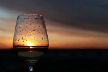 Glass of wine with water drops on background of sunset sky, sun and clouds are reflected in alcohol drink. Concept of celebration, home party, romantic dinner outdoors
