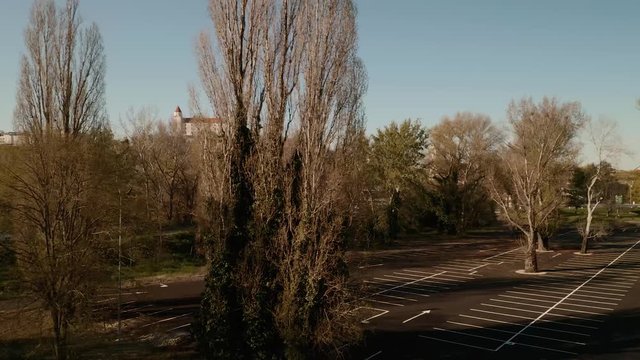 Aerial dolly shot of an empty parking lot with trees during the COVID-19 pandemic. The camera is moving from left to right revealing the Bratislava castle in the background behind the trees.