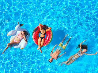 Aerial of friends having party in swimming pool with inflatable flamingo, swan, mattress. Happy young people relax at luxury resort on sunny day. View from above. Girls in bikini sunbathing in a sun.