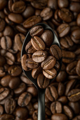Coffee beans in black metal spoon in the foreground over a background of coffee beans