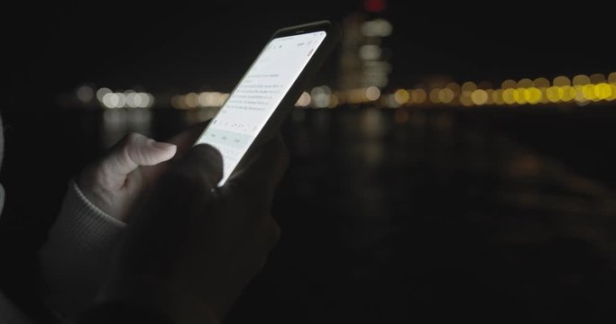 Female hands typing text message smartphone at night city background. Connecting and searching of social network. Blurred background and bokeh effects. Handheld close-up shot. 4K UHD