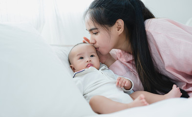 Asian mother kiss his child happily. In the bedroom with white curtains. Motherhood, family relationship, new born baby, love of mother to her son or daughter concept.