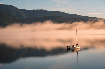 A lone sailboat is anchored in the remote bay of Deep Cove, North Vancouver. The mountains, blue sky and pink fog are reflected in the glass-like water at sunrise.