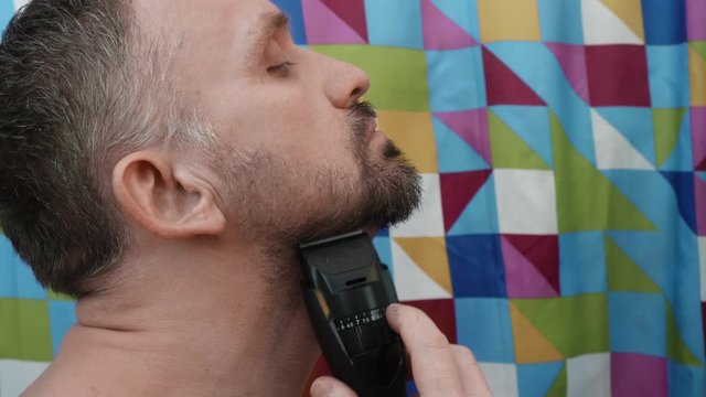 Mature man trims his beard with styler in front of mirror. Side view.
