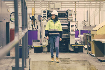Confident female printing worker walking at factory. Mature woman in yellow hard hat walking near printing machine and looking aside. Print manufacturing concept