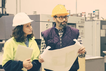 Two printing workers with blueprints. Focused employees holding blueprints and laptop and looking aside. Print manufacturing concept