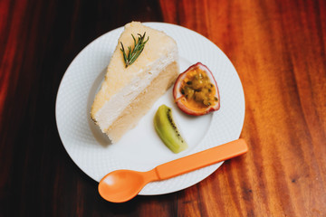 double fromage cheesecake served with sour fruit (passion fruit kiwi) in a white plate on a wooden table.Saw an orange spoon placed beside the plate