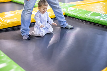 Little cute toddler girl in a dress jumping on a trampoline next to dad at a children's playground in a children's play center. blurred background