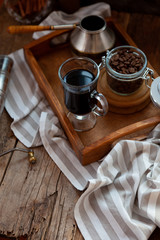 Obraz na płótnie Canvas A cup of coffee on a wooden tray. Turk and coffee beans. Cook breakfast at home. Still life with a drink in a glass with Cezve. Striped tablecloth on a wooden table. Cook at home.