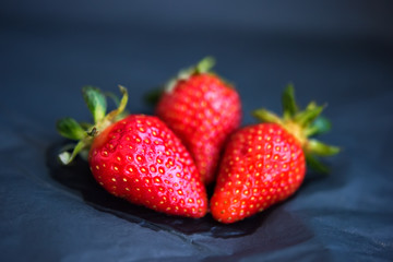 Three ripe fresh wet strawberries dark black crumpled paper background. Spring summer concept of vegetarian vegan food. Vitamins in fresh berries. Organic healthy fruits. selective focus. Defocused