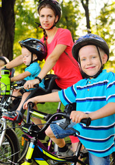 Naklejka premium a little boy in Bicycle gear and helmet is smiling while sitting on a Bicycle against the background of the Park, his mother and younger brother on bicycles.