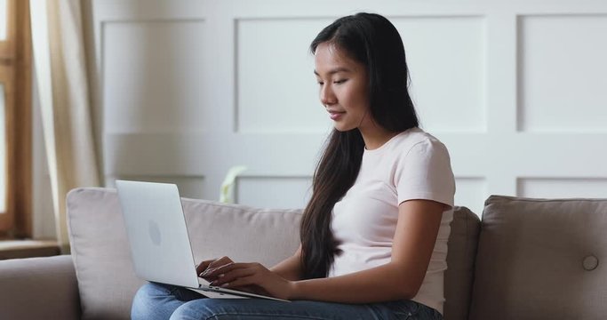Smiling focused asian ethnicity young woman sitting on couch, holding computer on lap, web surfing information. Happy professional vietnamese female freelancer working remotely from home on laptop.