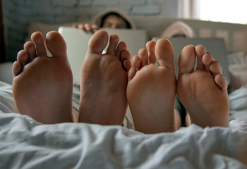 Fototapeta premium Bare feet in the foreground. Two teenagers are lying on a bed with laptops.