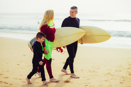 Family with surfboards walking on ocean coast. Side view of happy parents and little son in wetsuits holding hands and walking together on sandy beach. Surfing concept