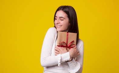 Fototapeta premium Cute girl is embracing a packed gift with eyes closed on yellow background.