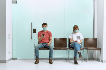 A guy and a girl in medical masks, sitting in a queue, and waiting for a doctor's appointment in...