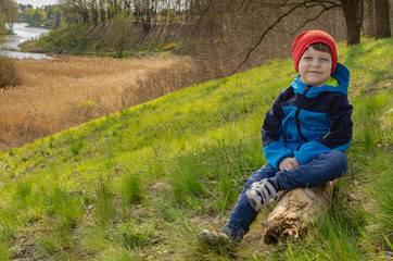 Cute boy a boy of four years old sits on a hill on a log and look at the lake. Family green tourism, walking with children outdoors