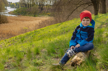 Cute boy a boy of four years old sits on a hill on a log and look at the lake. Family green tourism, walking with children outdoors