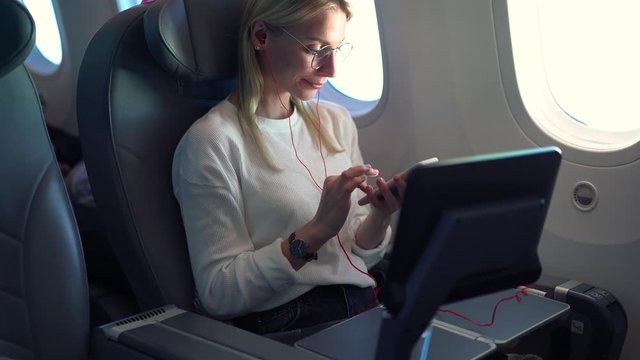 Attractive female in electronic headphones listening audio book with information about rest on board in airplane, woman tourist with cellular phone looking in window during flight in first class
