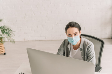 high angle view of freelancer in medical mask looking at camera while working at home