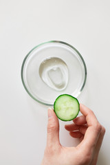 Woman hand holding slice of cucumber in hand and transparent cosmetic gel smudge in glass petri...