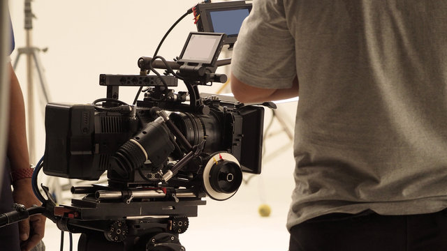 Behind the shooting production crew team and hd video camera and equipment in studio which includes big tripod, soft box light, monitors, lens for making online web film or movie or live broadcasting.