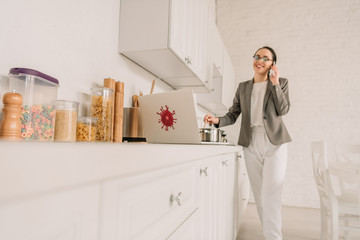 selective focus of businesswoman in a blazer over pajamas cooking and talking on smartphone in kitchen near laptop with coronavirus bacteria sticker