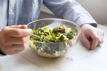 A man in a blue office shirt is sitting at a table with a bowl of salad. Cherry tomato salad with rucola, radicchio, lettuce, feta cheese, olive oil and balsamic dressing