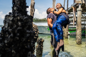 Loving couple in wetsuits under a wooden jetty.