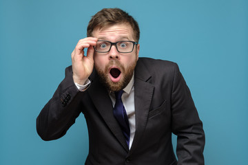 Surprised business man in a suit holds glasses in hand and in amazement opens mouth, looks with big eyes at the camera.