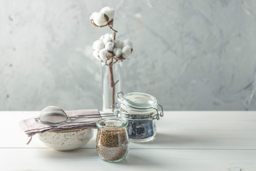 Coffee beans and instant coffee in glass jars, cotton flowers and kitchenware on white wooden table with grey concrete wall at background. Details of still life in the home interior. Cosy concept.
