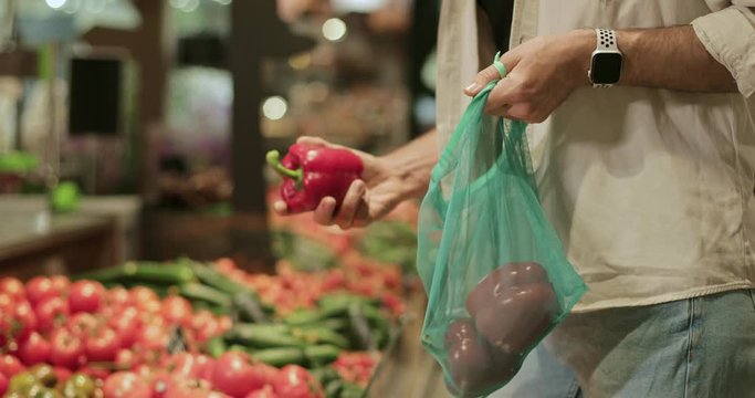 Crop view of man choosing and putting red pepper into reusable produce bag. Caucasian male person doing shopping in fruit and vegetables department in supermarket. Blurred background