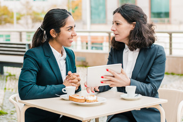Businesswomen drinking coffee and using tablet pc. Professional female coworkers in formal wear sitting at table and drinking coffee together in outdoor cafe. Coffee time concept