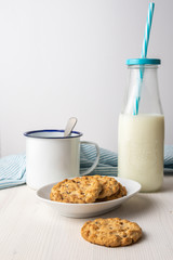 Close-up of chocolate chip cookie, white plate, cup with spoon, bottle of milk with blue straw and blue cloth, on table and white background