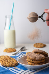 Close-up of colander with chocolate powder falling on white plate with chocolate chip cookies, spoon, bottle of milk, with selective focus, on blue napkin and white background, vertical