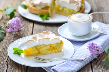 Leckerer Rhabarberkuchen mit Baiserhaube und Cappuccino auf einem Holztisch mit frühlingshafter mit Fliederdeko -  Delicious rhubarb cake with springlike lilac decoration