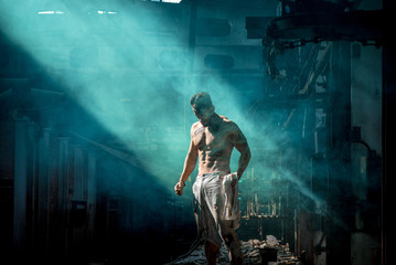 strong man against blue fog at factory exhausted stands, press and lateral abdominal muscles and pectoral muscles illuminated by light, place for text.