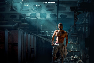 Fototapeta na wymiar sporty bodybuilder confidently stands in metal production plant, shines side light, man with press and biceps, blue smoke on backlight illuminated by light, place for text.
