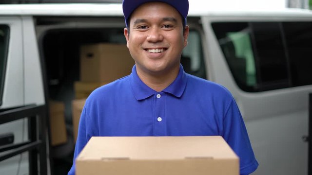 Corona Virus concept, Smiley blue delivery handsome asian man holding parcel cardboard box standing in front of the van. 4k resolution and Slow motion shot.
