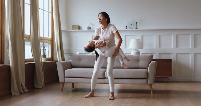 Emotional laughing young vietnamese mother lifting holding happy cute baby daughter, dancing barefoot in modern living room. Overjoyed mixed race asian family playing having fun together at home.